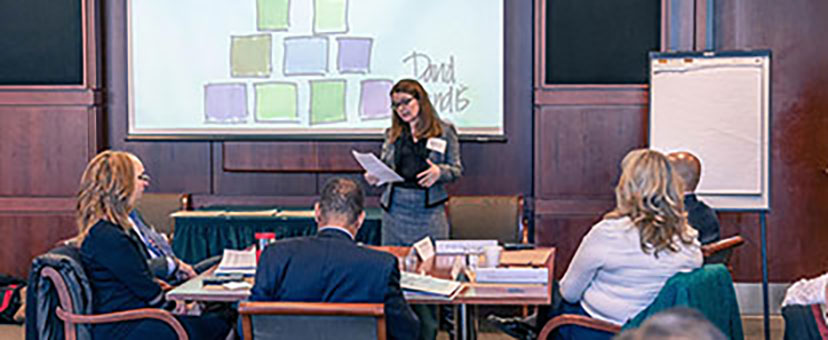 Speaker in front of a projection screen at an event with seated attendees Speaker in front of a projection screen at an event with seated attendees